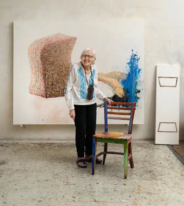Vera Klement with gray hair and glasses wearing a white and blue shirt, black pants and sandals stands in front of one of her paintings and rests her left hand on a simple chair that has been painted in many different colors.