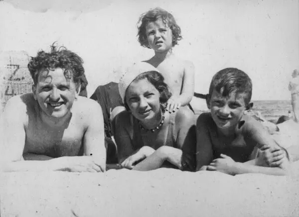 A black and white family portrait on what appears to be a beach. A man, a woman and a child are laying on their stomachs, propped up on their elbows on the sand. Another child sits on top of the woman's back.